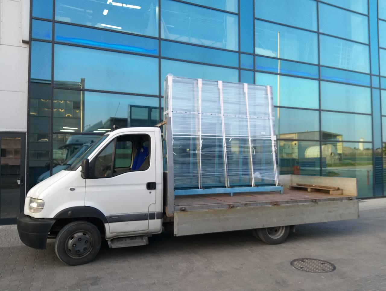 Biały bus z otwartą platformą załadowany pakietami szklanych paneli, zapakowanych w folię, na tle nowoczesnego budynku z niebieskimi szklanymi fasadami.