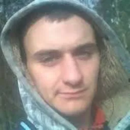 Portret mężczyzny w bluzie z kapturem, z widocznymi śladami zmęczenia na twarzy, na tle rozmytej zieleni drzew.