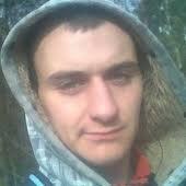 Portret mężczyzny w bluzie z kapturem, z widocznymi śladami zmęczenia na twarzy, na tle rozmytej zieleni drzew.