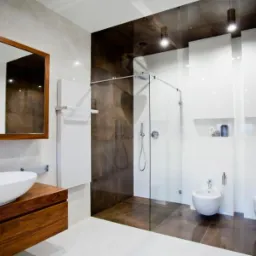 Nowoczesna łazienka z kabiną prysznicową typu walk-in, umywalką nablatową na drewnianej szafce, białymi szafkami wiszącymi, bidetem i toaletą podwieszaną. Ciemnobrązowe płytki imitujące marmur...