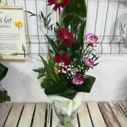 Kompozycja kwiatowa w szklanym wazonie z gerberami, goździkami i tulipanami, owinięta białym, ozdobnym papierem, na tle drewnianych desek i metalowej siatki z dekoracjami.