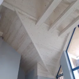 Drewniany sufit poddasza z widocznymi belkami i oknem, wykończony w jasnym odcieniu drewna, z fragmentem szarej ściany w dolnej części kadru.