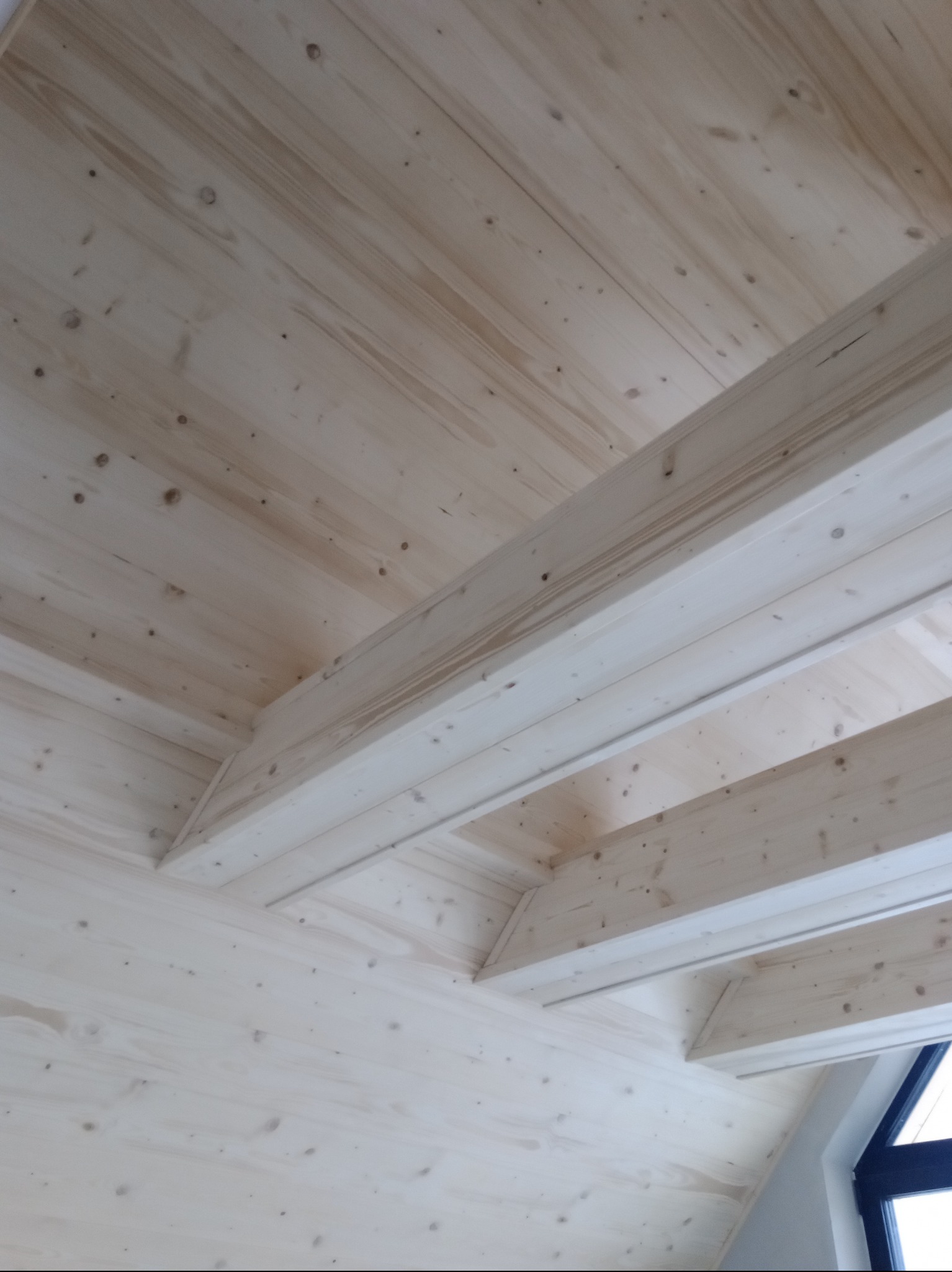 Jasna, drewniana boazeria sufitowa z widocznymi belkami konstrukcyjnymi, oświetlona naturalnym światłem wpadającym przez okno. Widok z dołu.