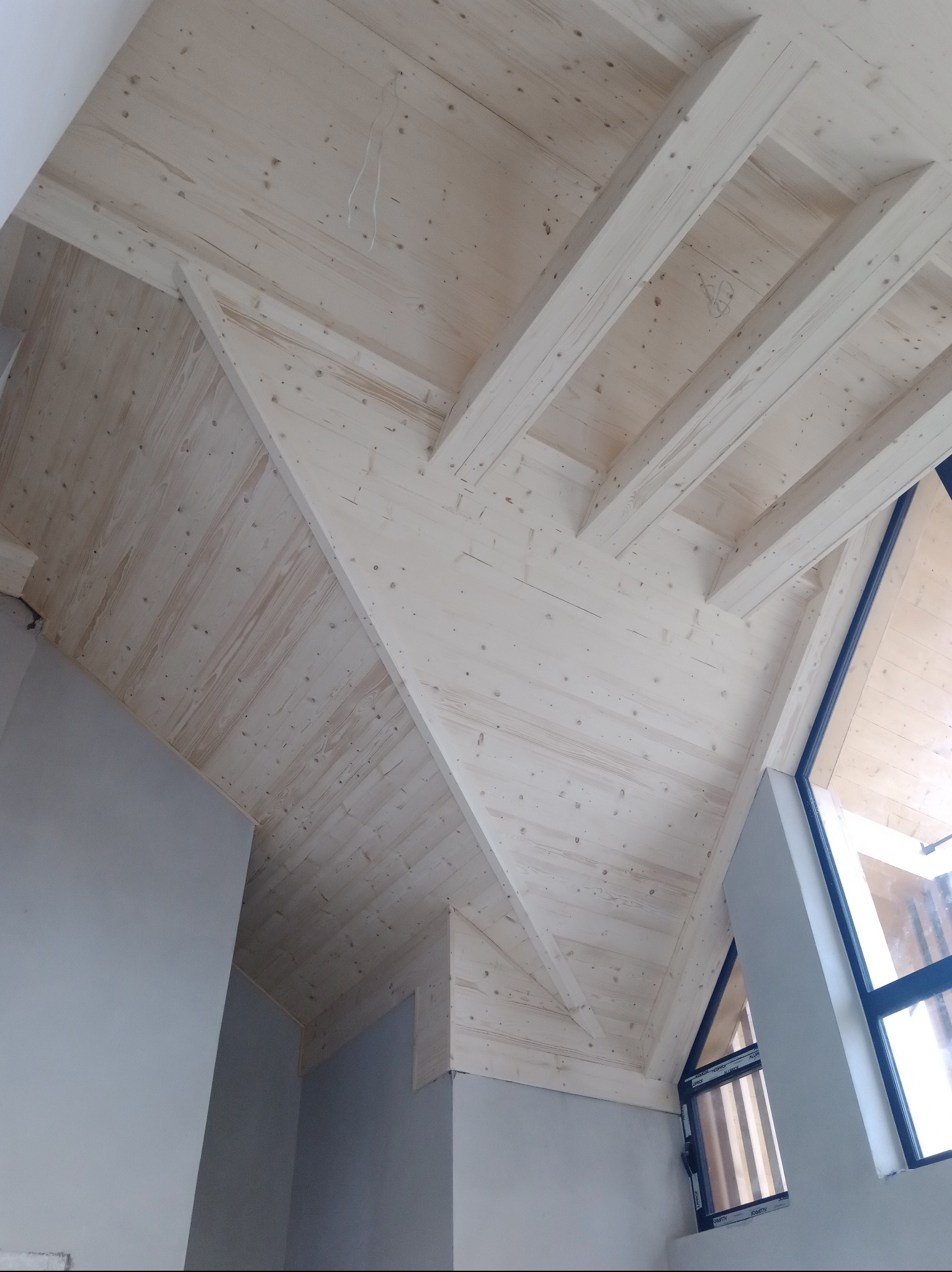 Drewniany sufit poddasza z widocznymi belkami i oknem, wykończony w jasnym odcieniu drewna, z fragmentem szarej ściany w dolnej części kadru.