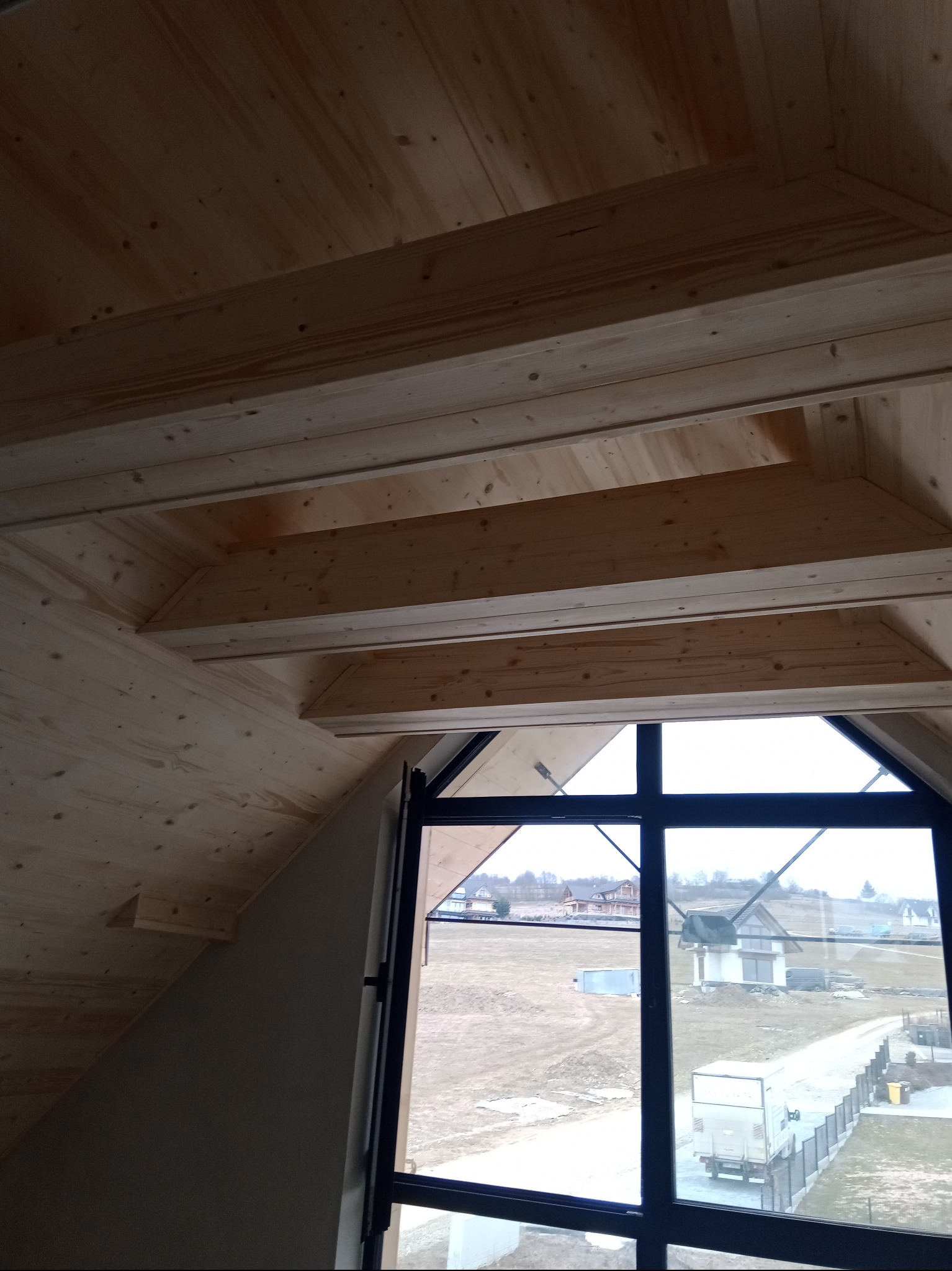 Drewniane wykończenie poddasza z widokiem na krajobraz przez okno o nietypowym kształcie. Widoczne belki stropowe i skosy. Naturalne światło dzienne.