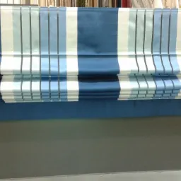 Niebiesko-białe rolety rzymskie w paski, częściowo zwinięte, prezentowane na tle szarej ściany w sklepie z tkaninami.