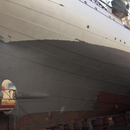 Sand Blast - Fragment kadłuba statku w doku, częściowo pokryty świeżą, szarą farbą antykorozyjną, widoczne ślady wcześniejszych powłok i konstrukcja nitowana.