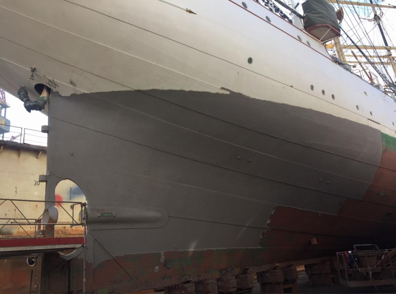 Fragment kadłuba statku w doku, częściowo pokryty świeżą, szarą farbą antykorozyjną, widoczne ślady wcześniejszych powłok i konstrukcja nitowana.