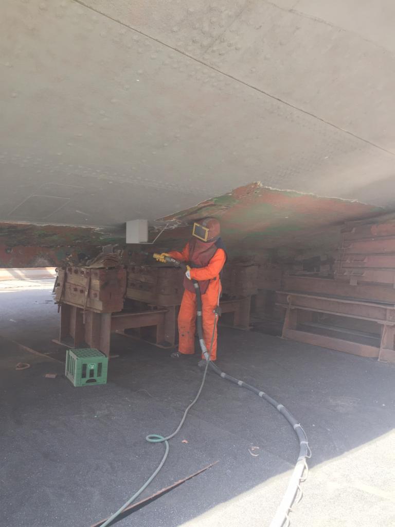 Pracownik w kombinezonie ochronnym i masce piaskuje metalową konstrukcję, usuwając starą farbę i rdzę, widoczny wąż doprowadzający materiał ścierny i zielona skrzynka w tle.