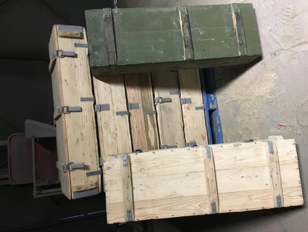 Stos drewnianych skrzyń transportowych, niektóre z metalowymi okuciami, ułożonych na niebieskiej palecie, w magazynie o betonowej podłodze.