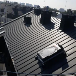 Nowoczesny, grafitowy dach z blachy trapezowej z czterema kominami i oknem dachowym, widok z góry na tle zabudowy miejskiej w oddali.