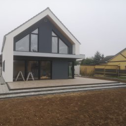 DAS-BUD - Nowoczesny dom z tarasem wyłożonym jasnymi płytami, otoczony świeżo przygotowanym terenem pod trawnik, widoczne schody z kostki brukowej i dwie drabiny opierające się o ścianę.