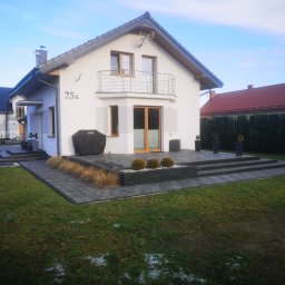 DAS-BUD - Nowoczesny dom jednorodzinny z jasną elewacją i ciemną kostką brukową na podjeździe oraz schodach wejściowych, widoczne elementy zagospodarowania ogrodu z niskimi krzewami i trawami ozdobnymi.