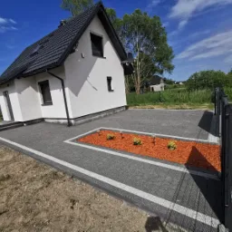 Nowoczesny dom z szarą kostką brukową, obrzeżami z białymi paskami i rabatą z kory oraz niskimi krzewami i lampkami ogrodowymi. Metalowe ogrodzenie w tle.