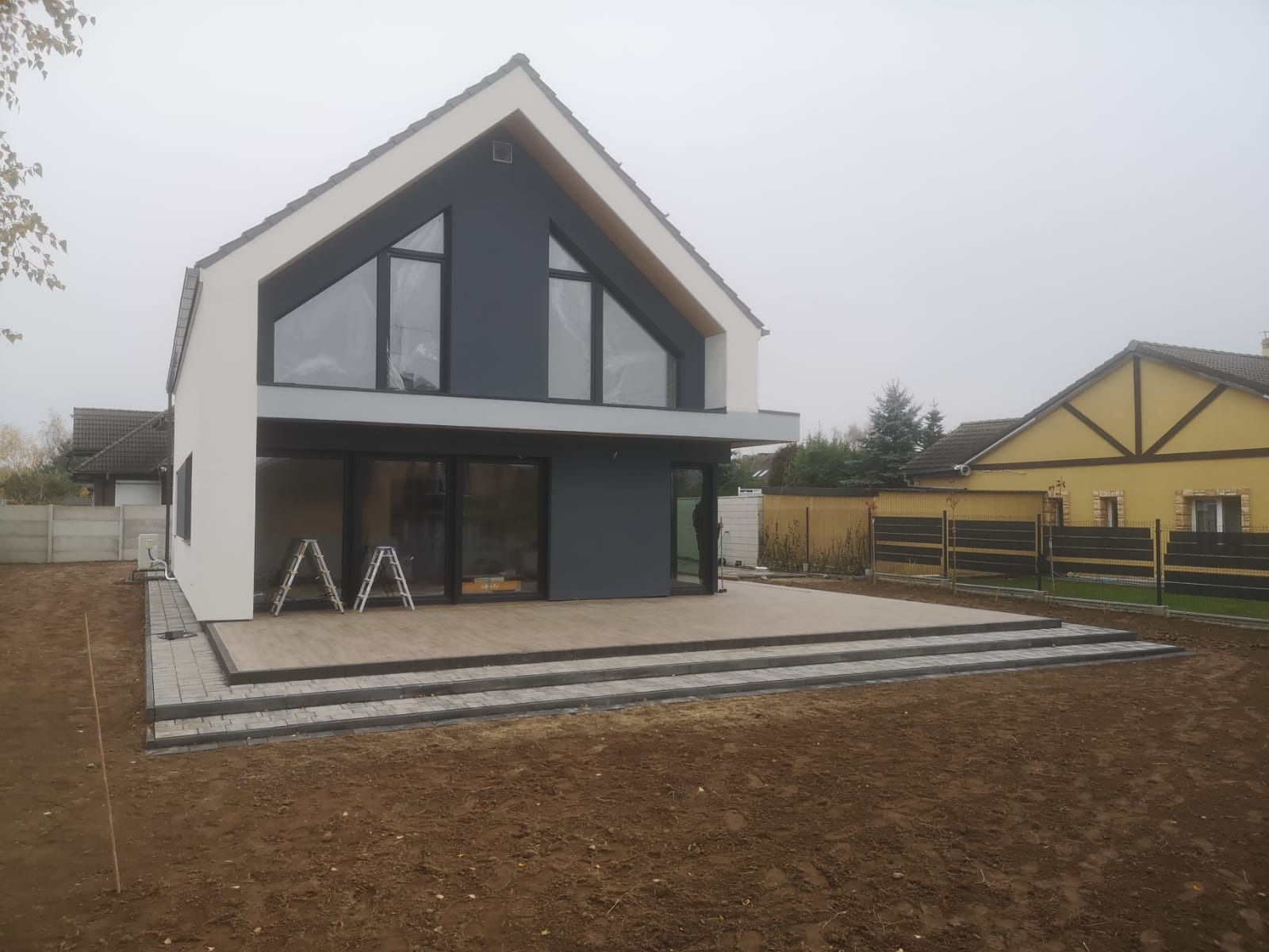 Nowoczesny dom z tarasem wyłożonym jasnymi płytami, otoczony świeżo przygotowanym terenem pod trawnik, widoczne schody z kostki brukowej i dwie drabiny opierające się o ścianę.