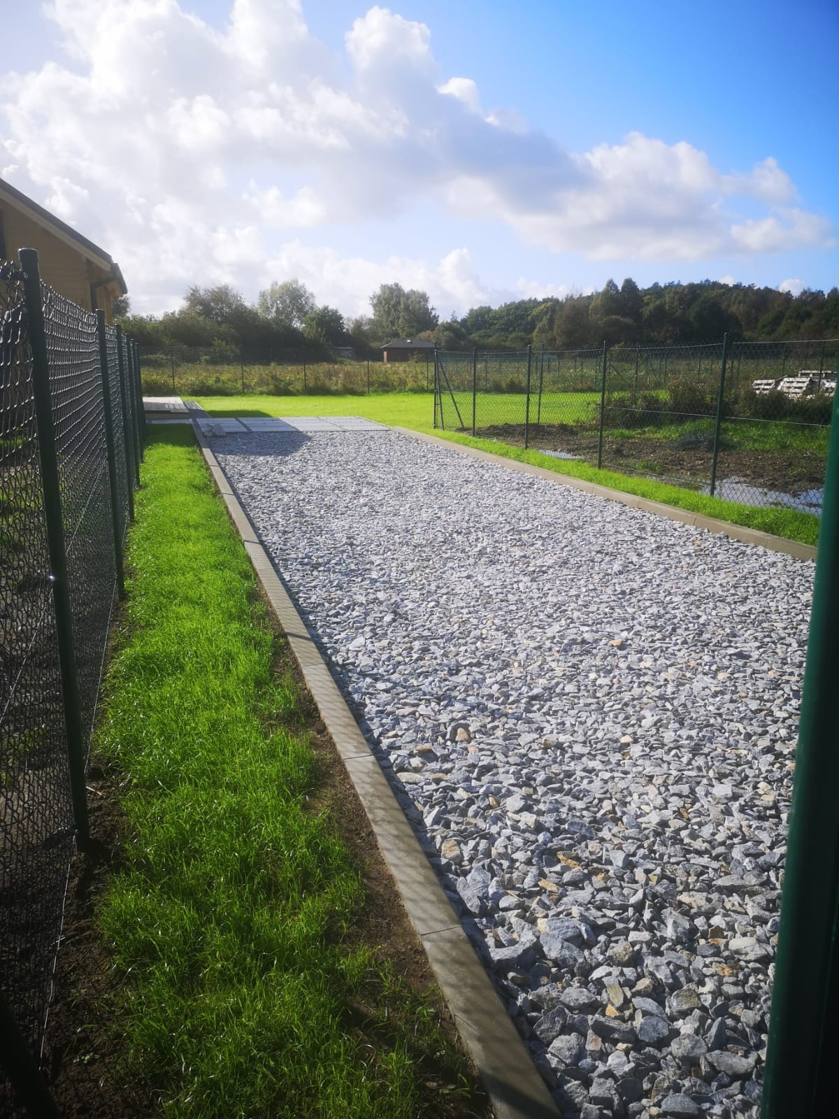 Długa, utwardzona żwirem droga dojazdowa z betonowymi obrzeżami, widok zza ogrodzenia, w tle zielona trawa i drzewa pod błękitnym niebem z chmurami.
