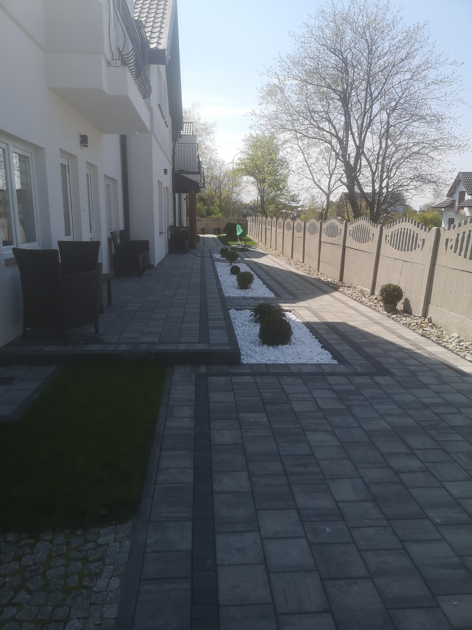 Ułożona szara kostka brukowa tworząca ścieżkę wzdłuż białego domu z balkonem, oddzielona pasami białych kamieni i zieleni, zakończona betonowym ogrodzeniem.