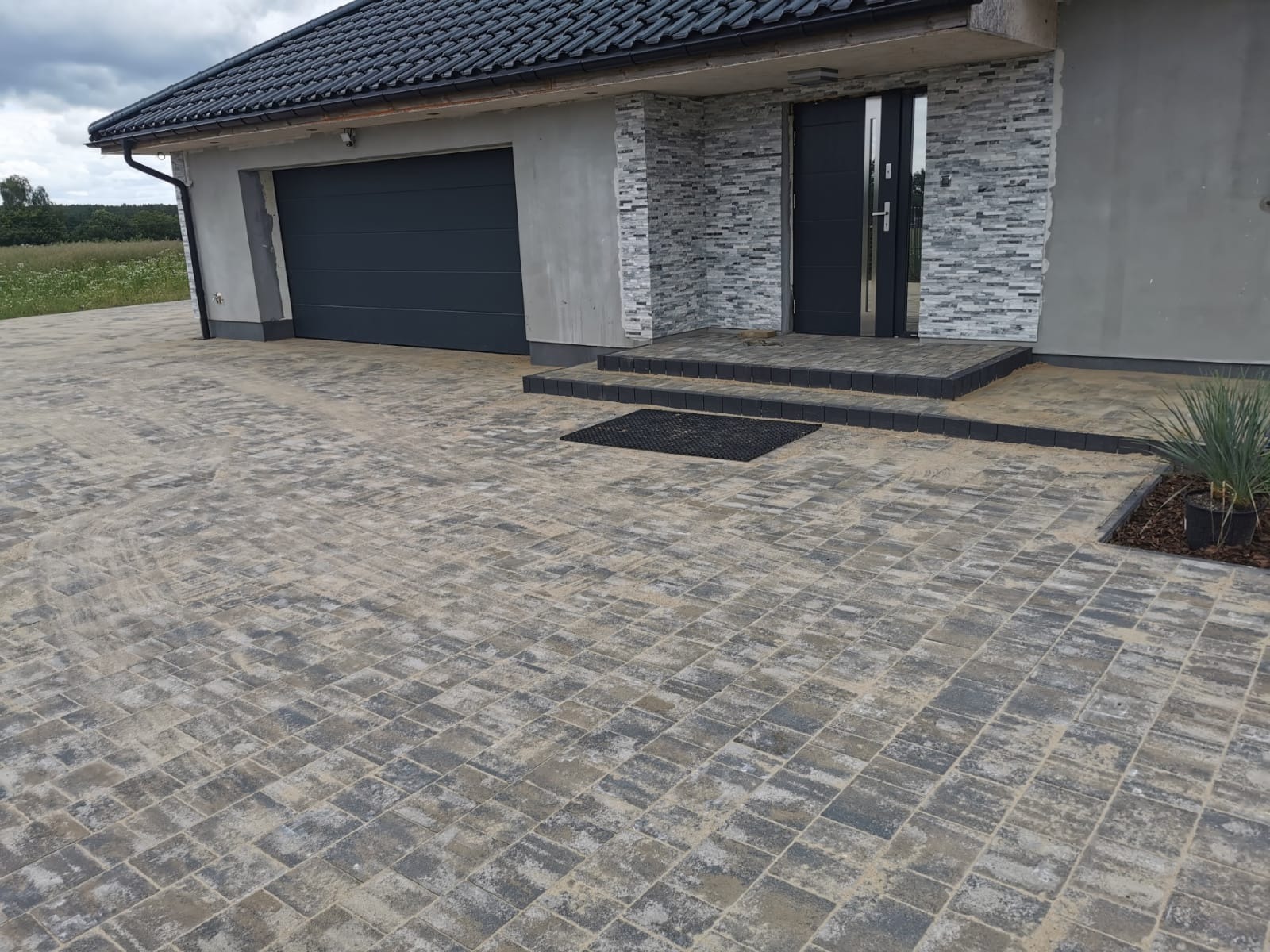 Nowo ułożona kostka brukowa w odcieniach szarości i beżu prowadząca do garażu z automatyczną bramą i wejścia do domu z czarnymi drzwiami i okładziną z szarego kamienia.