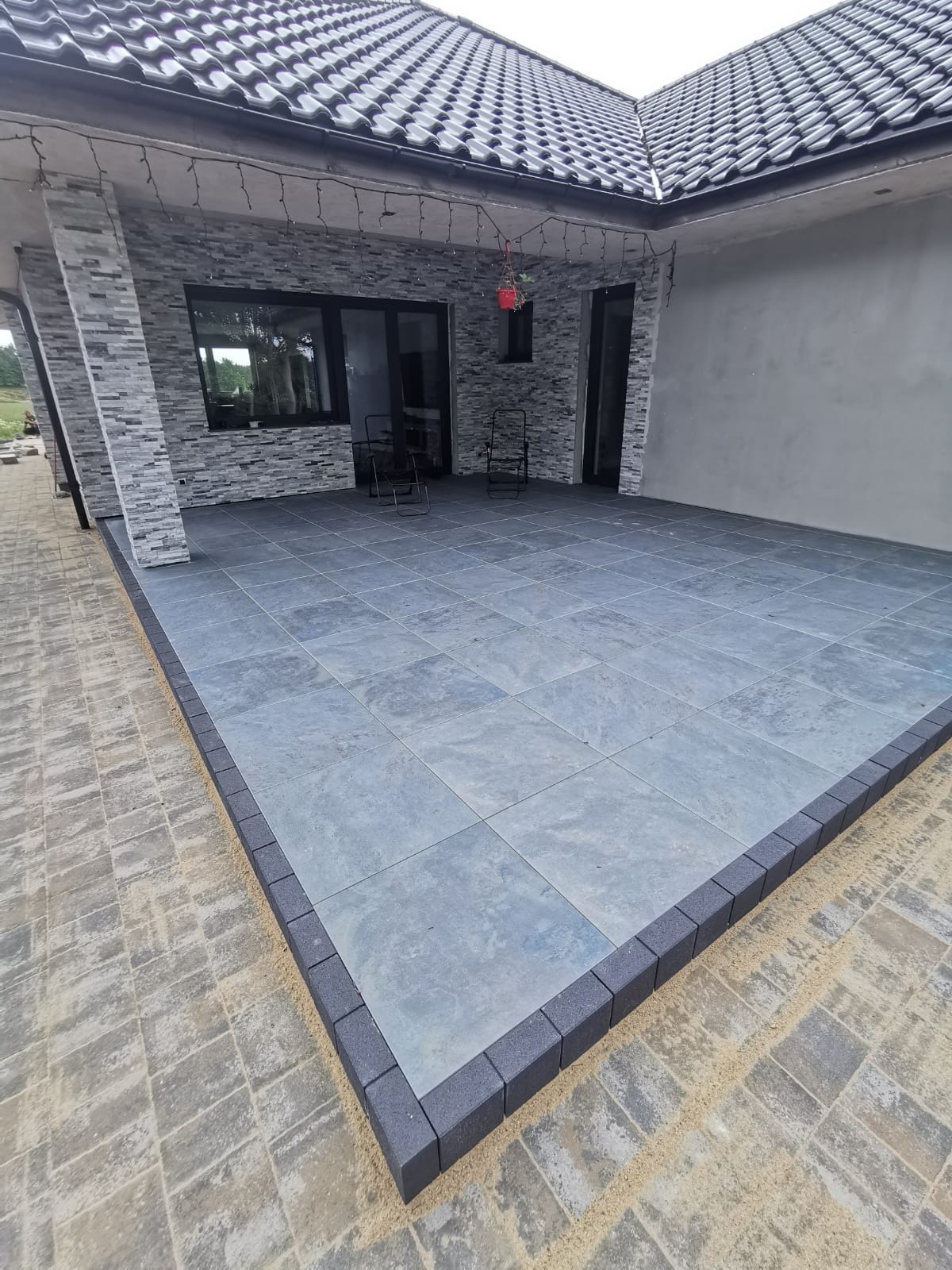 Nowoczesny taras wyłożony szarą, kwadratową płytą z ciemnoszarą obwódką, przylegający do domu z elewacją z szarej cegły i ciemnym dachem z dachówki.