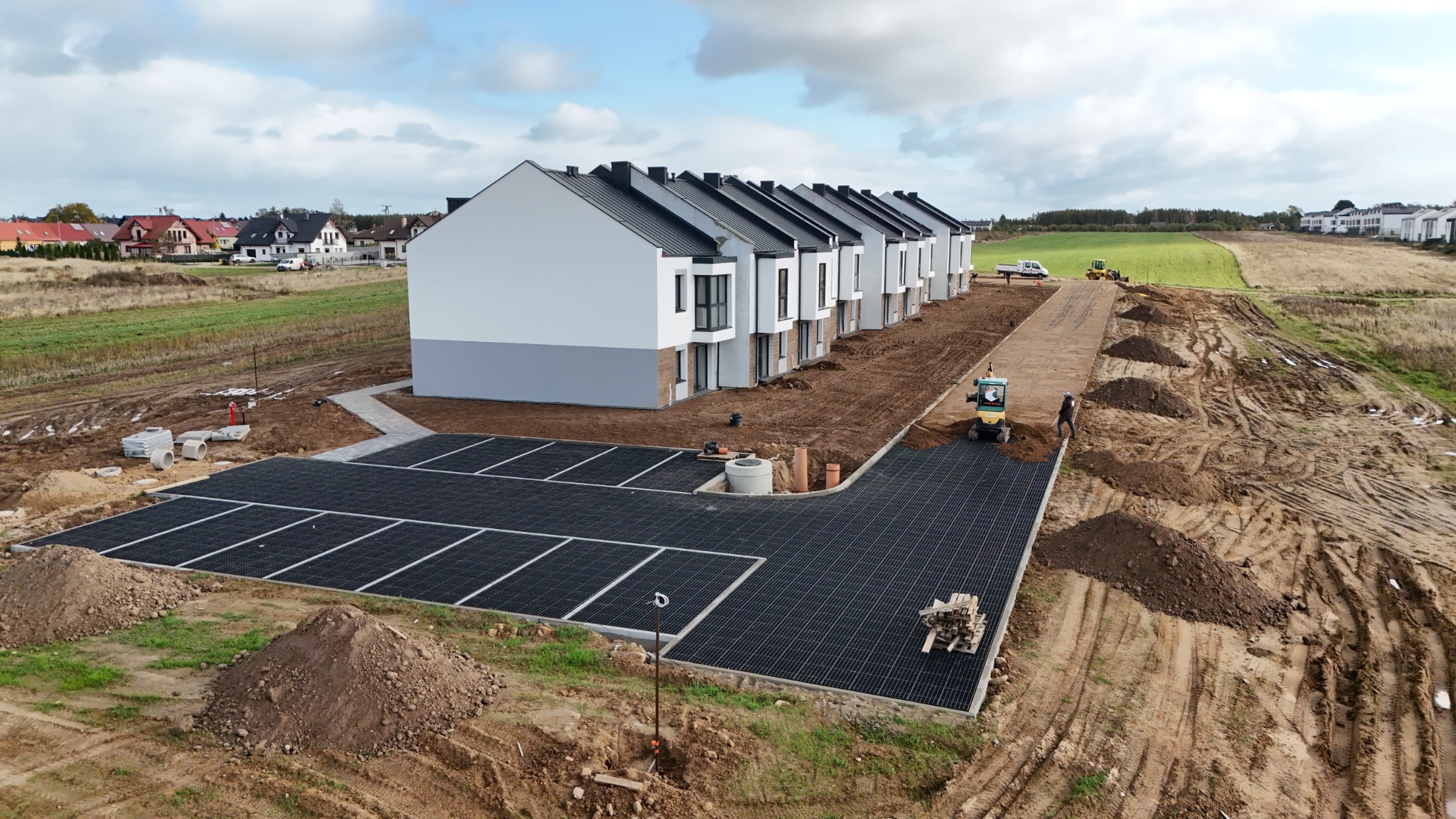 Widok z góry na nowo wybudowane szeregówki z wykonanym parkingiem z ażurowych płyt, teren przygotowywany pod drogę dojazdową z użyciem mini koparki.
