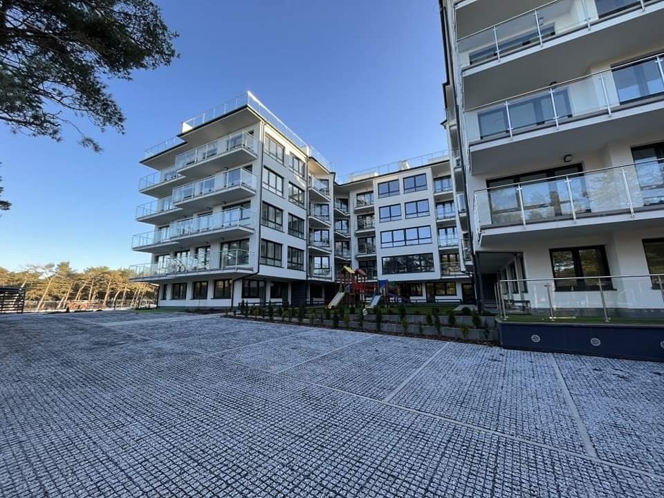 Nowoczesny apartamentowiec z balkonami i szarą, ażurową nawierzchnią parkingu, widok z poziomu gruntu w słoneczny dzień.