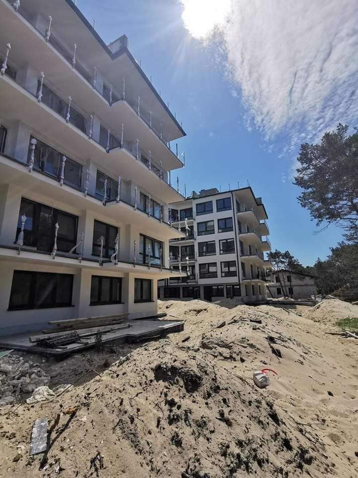 Nowoczesny budynek apartamentowy w trakcie budowy, widok na fasadę z balkonami i teren wokół budynku z piaskiem i elementami budowlanymi, błękitne niebo z chmurami w tle.