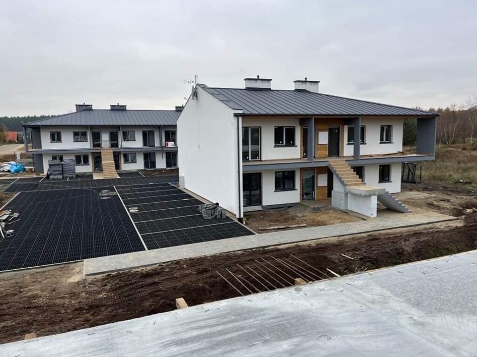 Nowe dwukondygnacyjne domy szeregowe z zewnętrznymi schodami na piętro i brukowanym parkingiem, widok z góry na plac budowy.