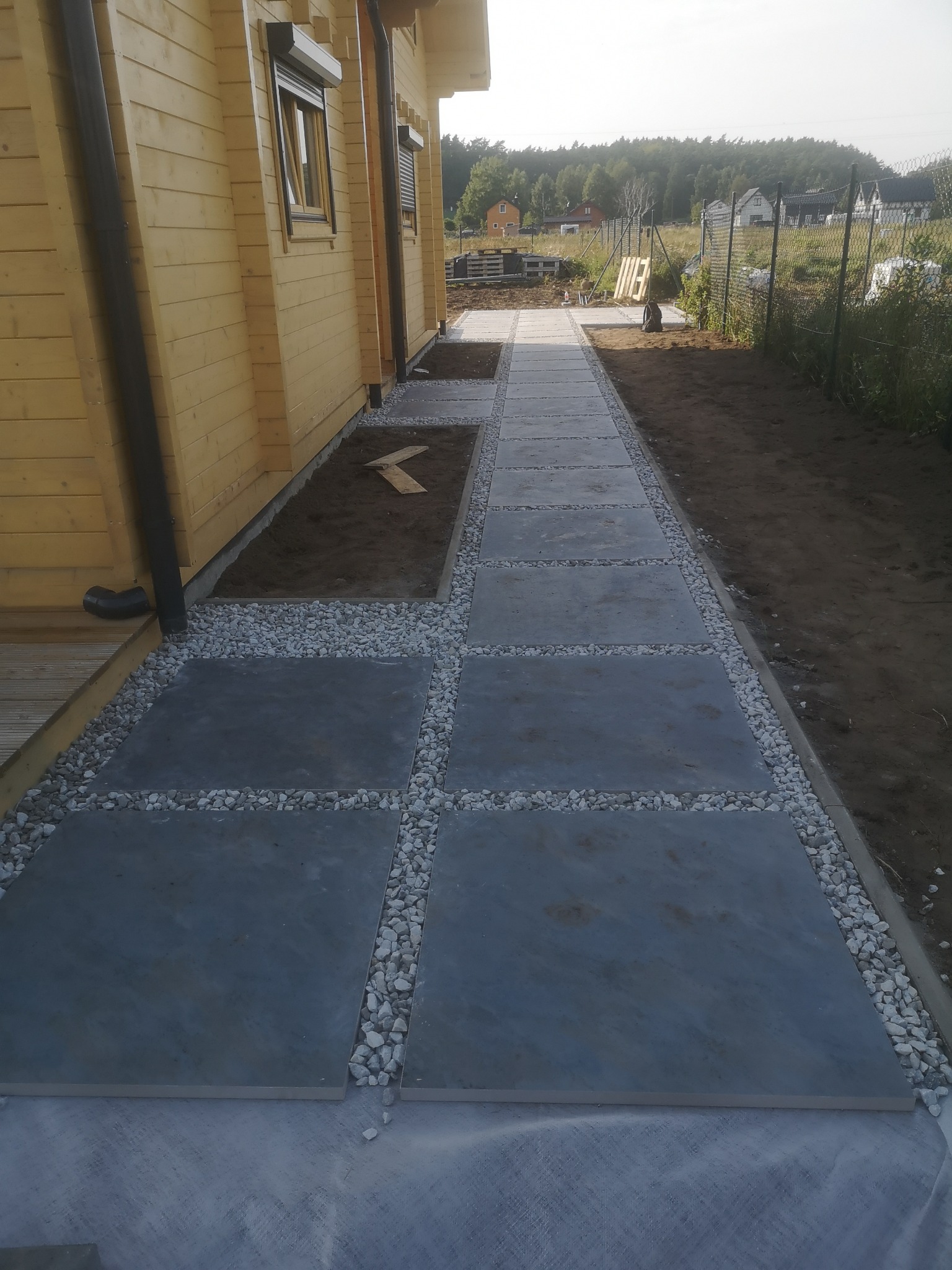 Ścieżka z szarych płyt ułożona w ogrodzie obok drewnianego domu, z białym żwirem między płytami i wzdłuż krawędzi, widoczna ziemia przygotowana pod kolejne płyty.