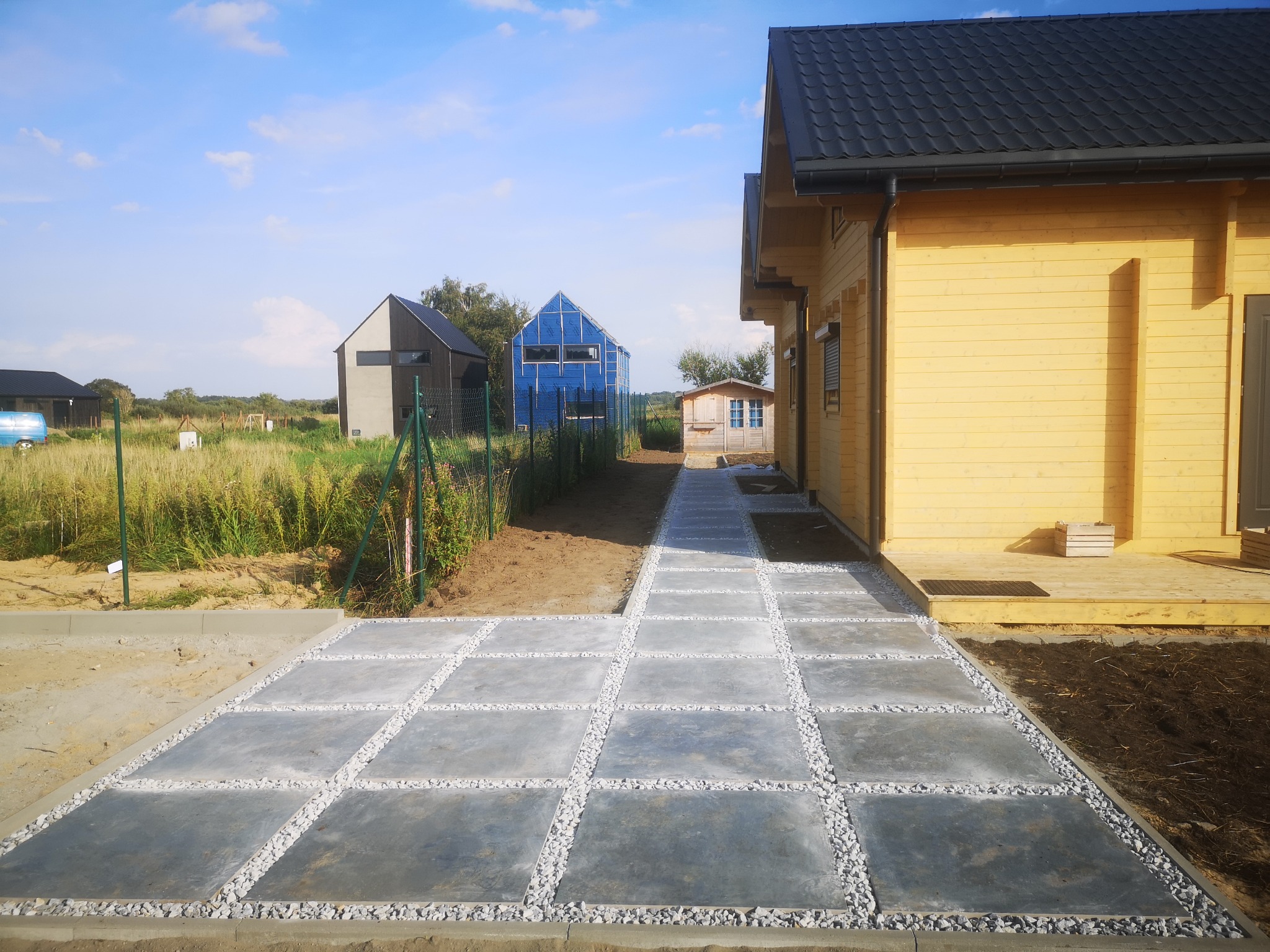 Ułożona ścieżka z kwadratowych płyt betonowych oddzielonych białym grysem, prowadząca do drewnianego domu z czarnym dachem; w tle widoczne inne budynki i ogrodzenie.