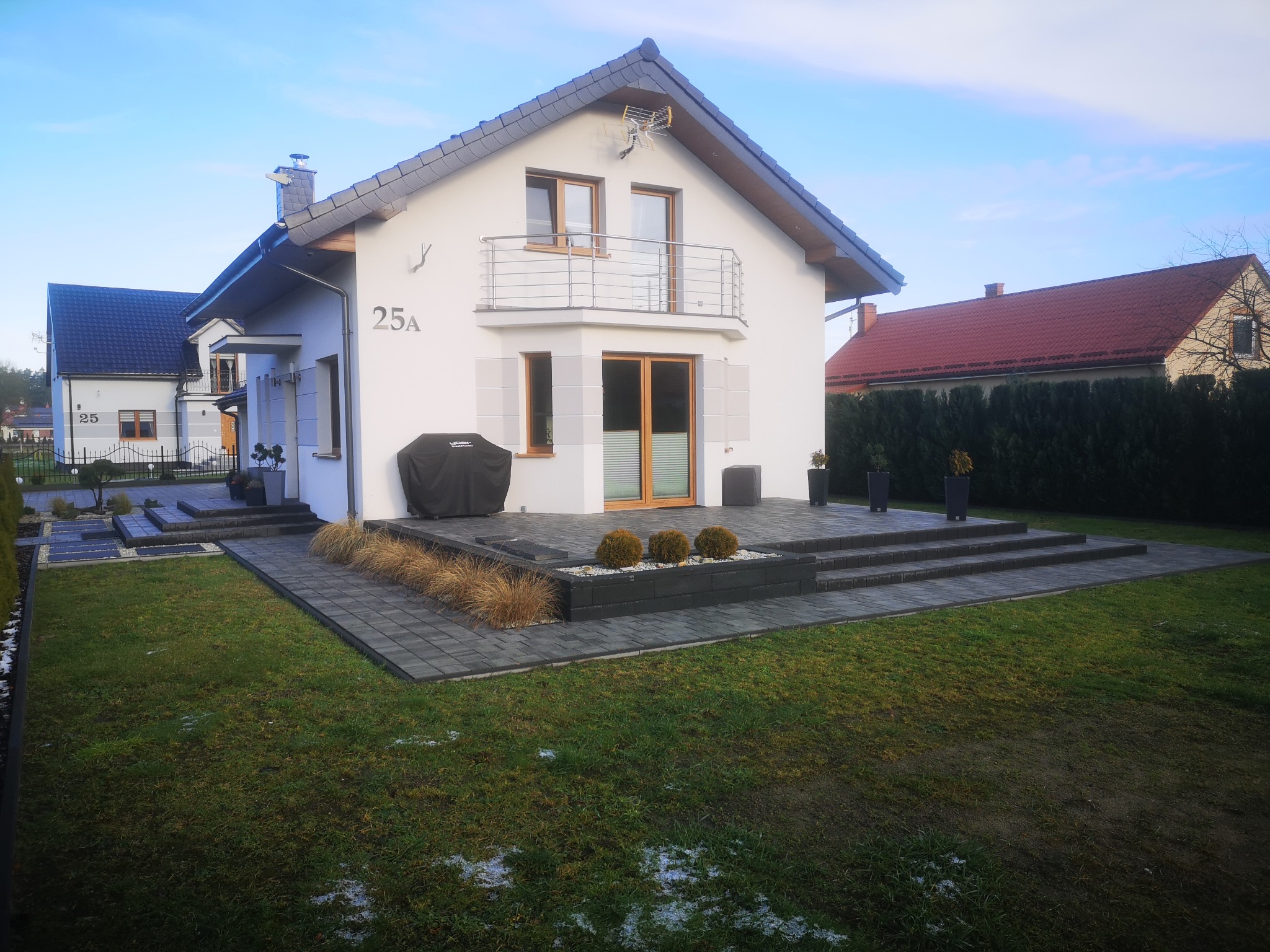 Nowoczesny dom jednorodzinny z jasną elewacją i ciemną kostką brukową na podjeździe oraz schodach wejściowych, widoczne elementy zagospodarowania ogrodu z niskimi krzewami i trawami ozdobnymi.