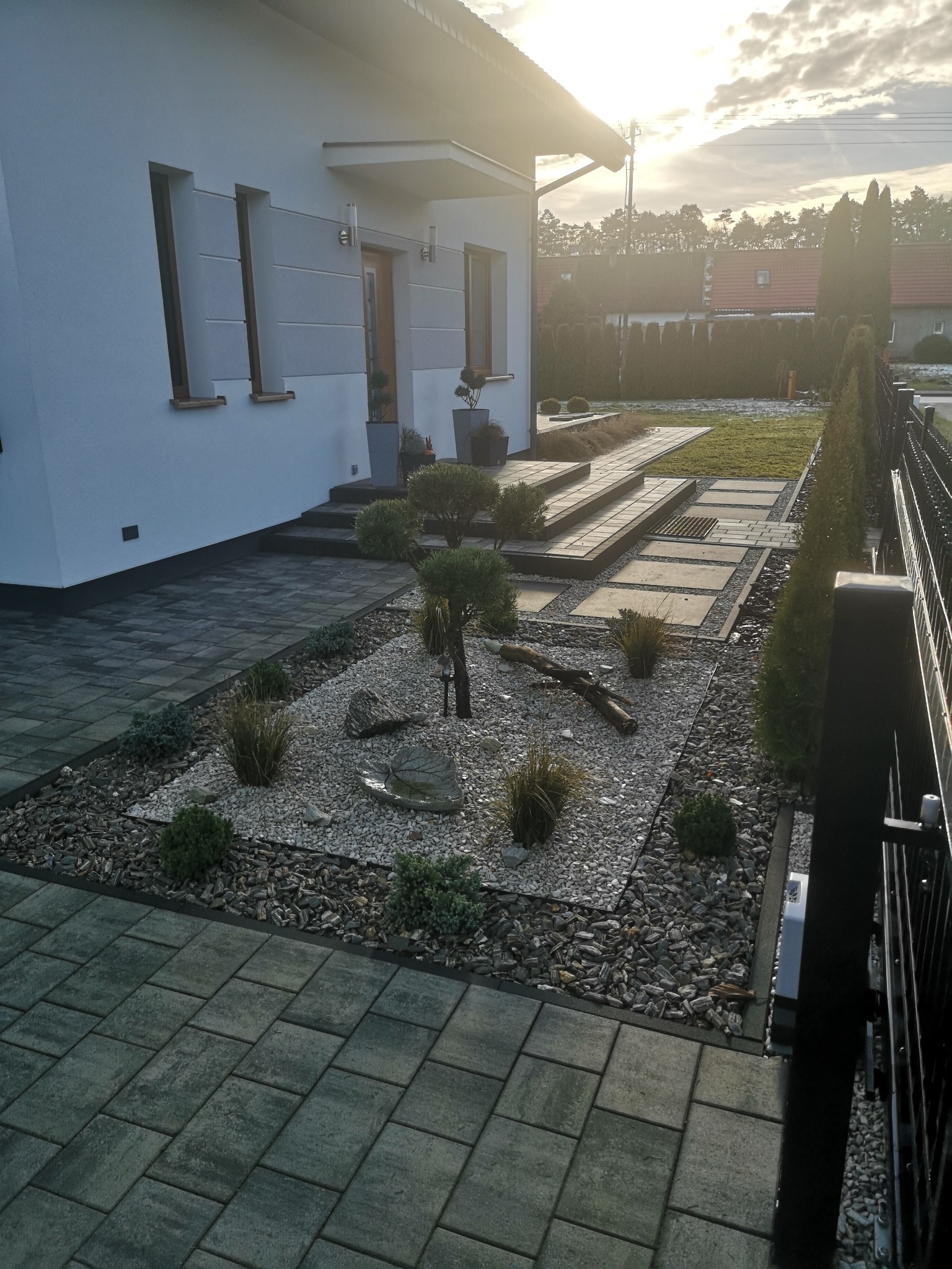 Nowoczesny ogród przed domem z kostką brukową, kamieniami ozdobnymi i niską roślinnością, widok na elewację budynku i wschodzące słońce.