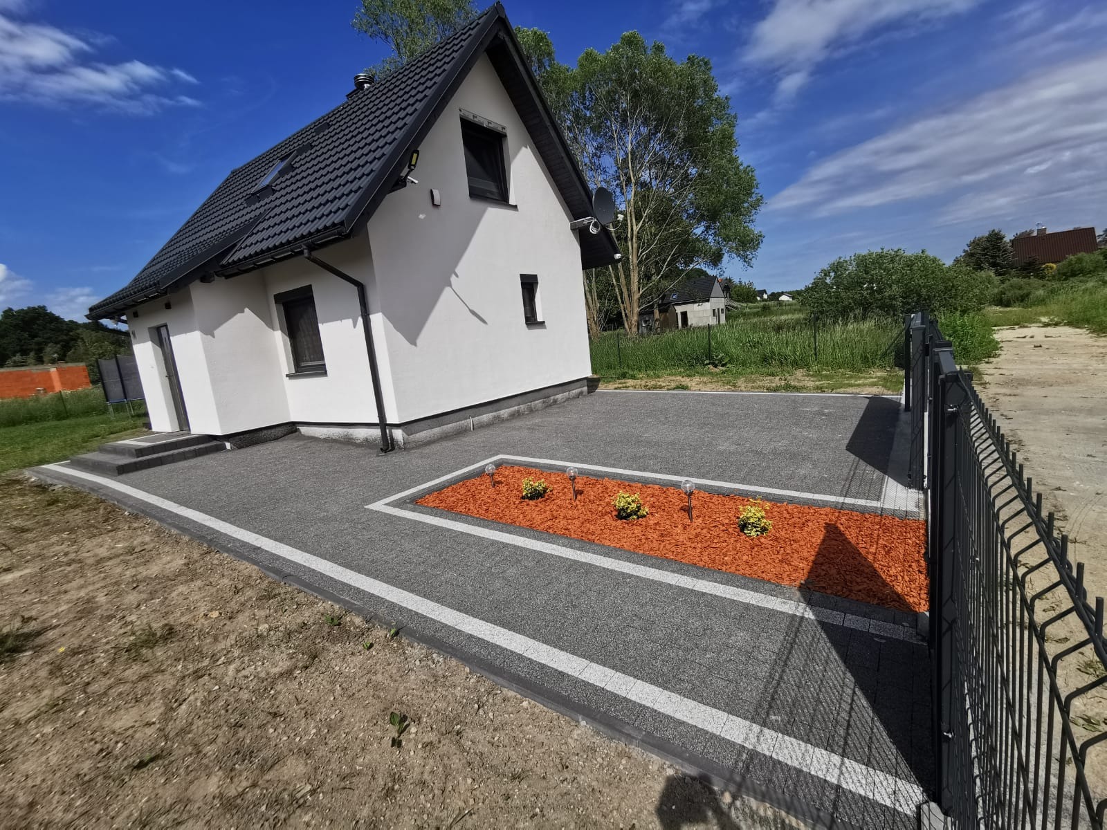 Nowoczesny dom z szarą kostką brukową, obrzeżami z białymi paskami i rabatą z kory oraz niskimi krzewami i lampkami ogrodowymi. Metalowe ogrodzenie w tle.