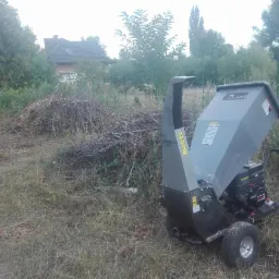Profesjonalny rozdrabniacz do gałęzi marki Tiler na trawiastym terenie, z widocznymi stertami pociętych gałęzi w tle, sugerujący prace porządkowe w ogrodzie.