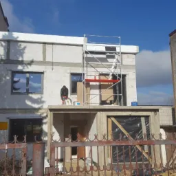 Budynek w trakcie ocieplania z widocznym pracownikiem używającym szlifierki do wyrównywania powierzchni, rusztowaniem i częściowo zamontowanymi oknami, sąsiedni budynek w tle.