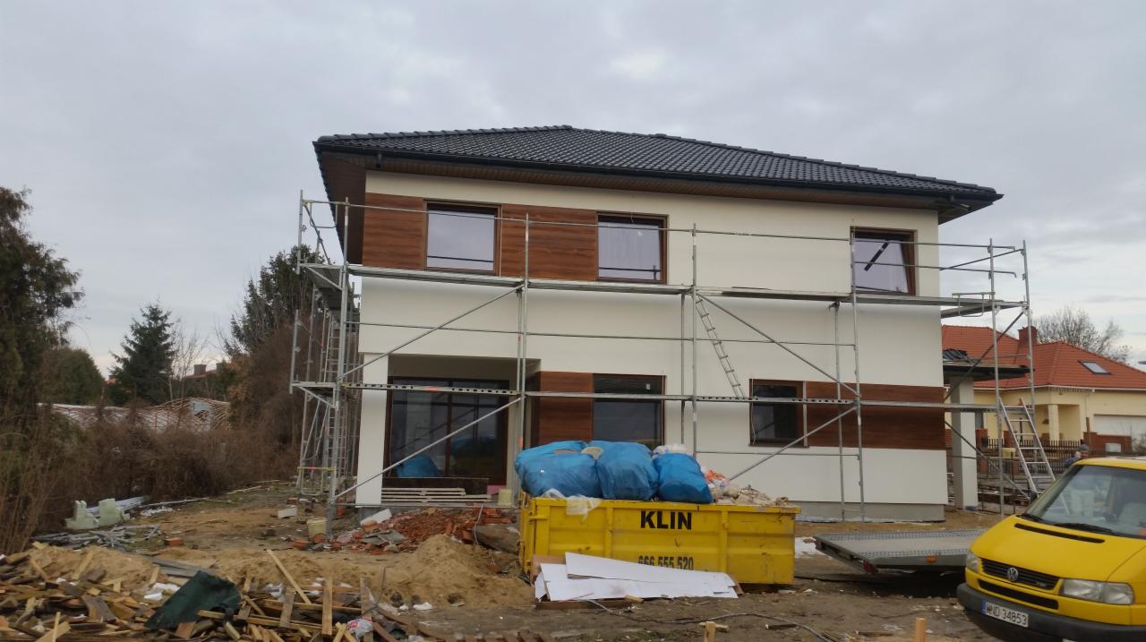 Dwukondygnacyjny dom w trakcie wykańczania elewacji, z widocznymi rusztowaniami, częściowo otynkowany na biało z poziomymi pasami z drewnopodobnych paneli, żółty kontener na gruz i żółty samochód...