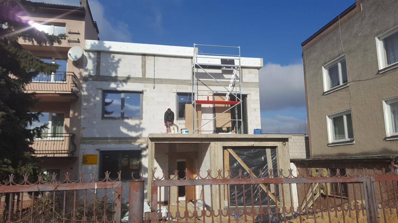 Budynek w trakcie ocieplania z widocznym pracownikiem używającym szlifierki do wyrównywania powierzchni, rusztowaniem i częściowo zamontowanymi oknami, sąsiedni budynek w tle.