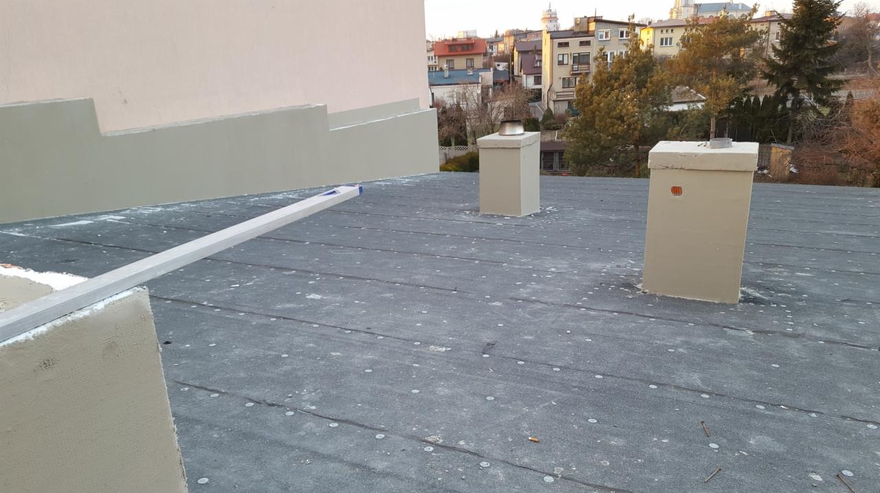 Prace na dachu w Szydłowcu: świeżo otynkowane kominy i fragment ściany budynku z widocznym poziomem, na tle zabudowy miejskiej.