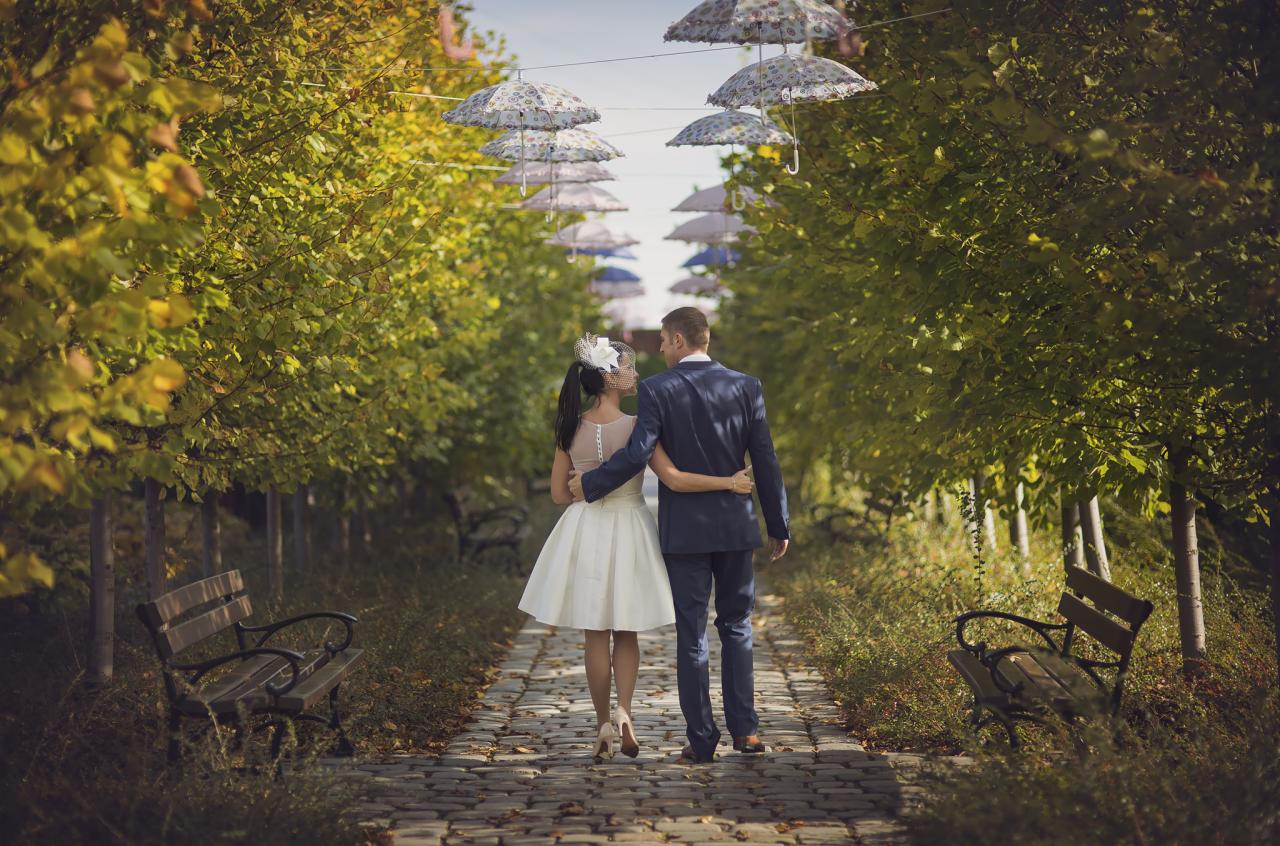 Para młoda, ubrana w eleganckie stroje, spaceruje aleją z drzew, nad którą zawieszone są dekoracyjne parasolki, w słoneczny dzień.