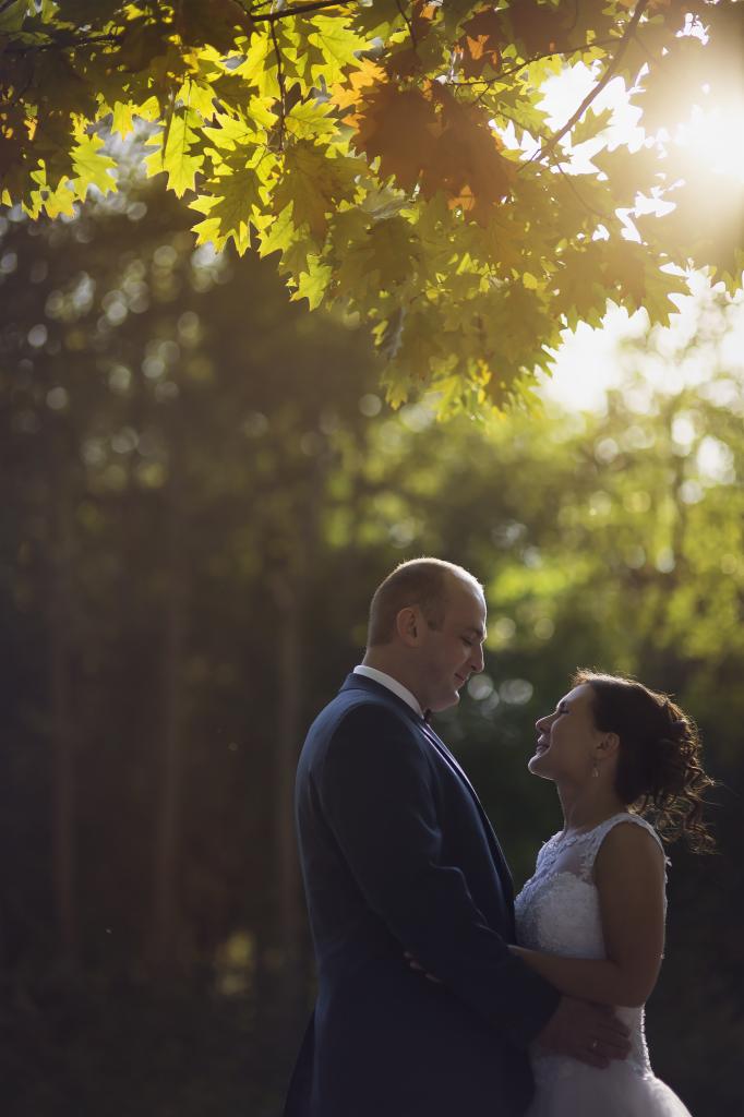 Para młoda wpatrzona w siebie pod drzewem z promieniami słońca przebijającymi przez liście, on w granatowym garniturze, ona w białej sukni ślubnej.