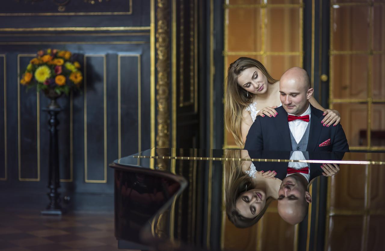 Elegancka para młoda: panna młoda obejmuje pana młodego stojącego przy fortepianie, widoczne odbicie w lakierowanej powierzchni instrumentu, w tle dekoracyjne ściany i bukiet kwiatów.