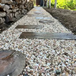 Ścieżka w ogrodzie wysypana drobnym, białym żwirem z drewnianymi i okrągłymi płytami chodnikowymi, obok ułożone drewno na opał.