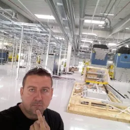 Instalacja przemysłowa w trakcie budowy z widoczną konstrukcją wsporczą pod sufitem, kanałami wentylacyjnymi i dużymi niebieskimi urządzeniami, z mężczyzną w czarnej koszulce robiącym selfie...