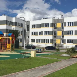 Nowoczesny, trzypiętrowy budynek mieszkalny z placem zabaw, elementami elewacji w kolorze białym, szarym i żółtym, z widocznym placem zabaw i zaparkowanymi samochodami.