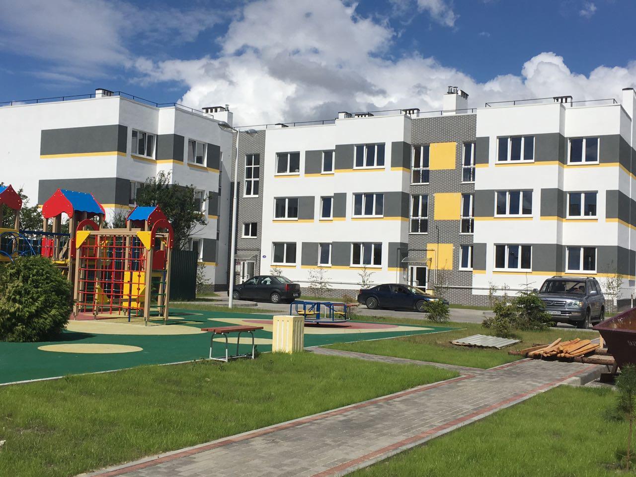 Nowoczesny, trzypiętrowy budynek mieszkalny z placem zabaw, elementami elewacji w kolorze białym, szarym i żółtym, z widocznym placem zabaw i zaparkowanymi samochodami.