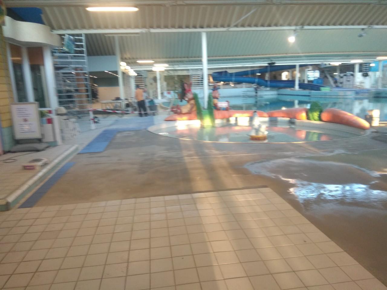 Wnętrze basenu w trakcie prac remontowych, widoczne płytki podłogowe, instalacje zabawne w kształcie marchwi i zjeżdżalnie w tle.