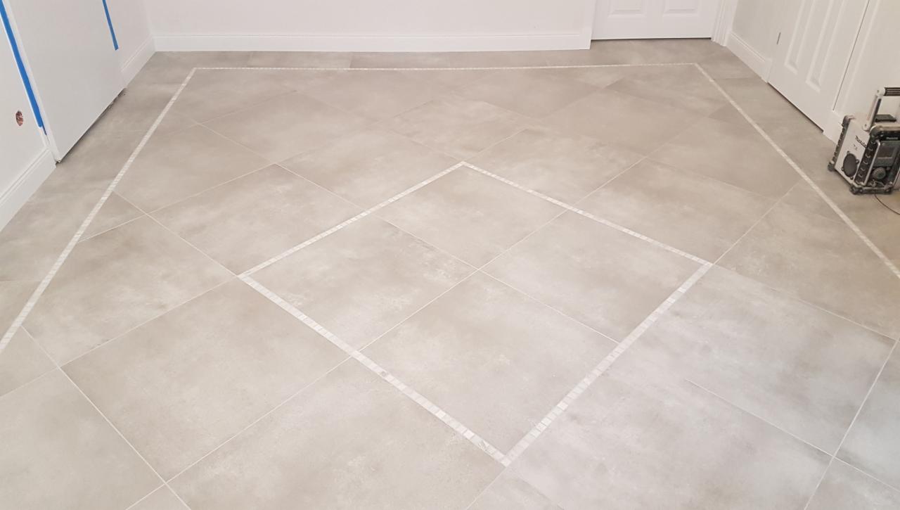 Beżowe płytki podłogowe ułożone pod kątem 45 stopni, z dekoracyjną ramką z mozaiki tworzącą romb na środku i obramowanie przy ścianach. W tle widoczne białe drzwi i sprzęt budowlany.