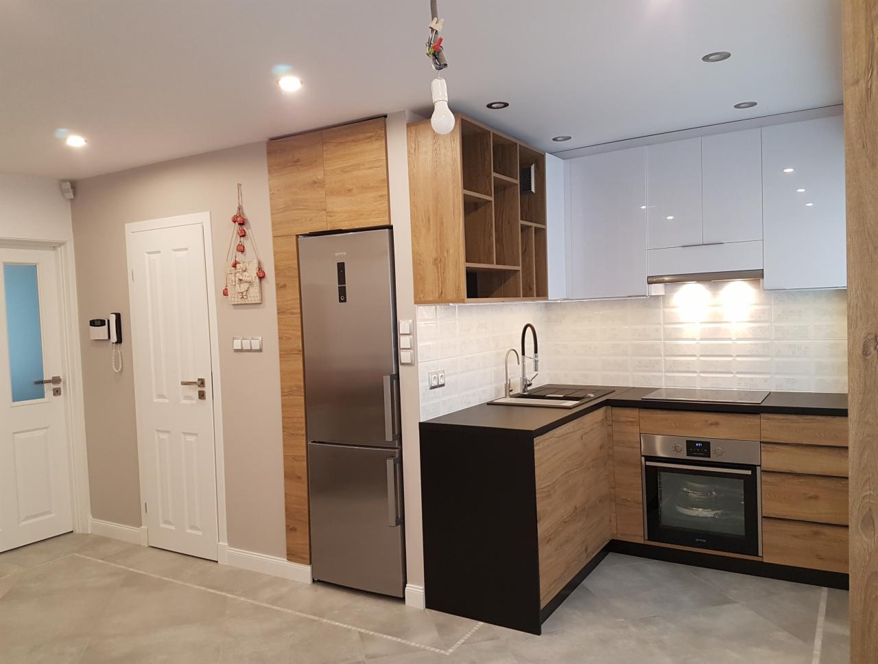 Nowoczesna kuchnia z drewnianymi i białymi szafkami, czarnym blatem, srebrną lodówką i szarą podłogą, oświetlona punktowymi lampami sufitowymi.