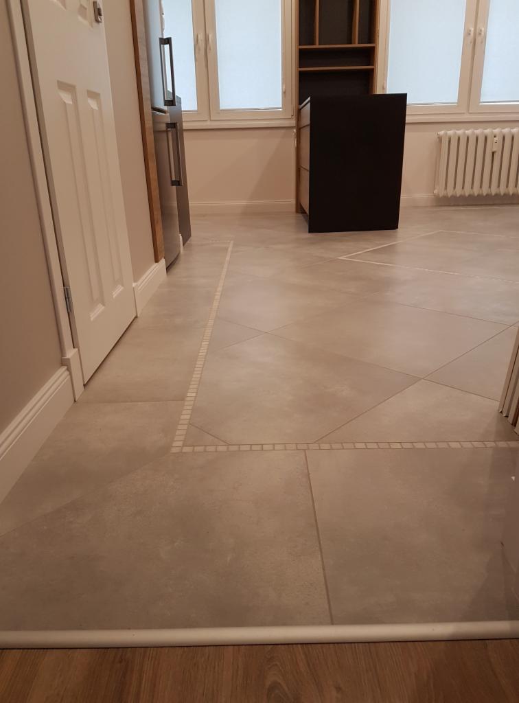 Beżowe płytki podłogowe ułożone w romby, oddzielone mozaiką, widok na fragment kuchni z meblami i oknem.