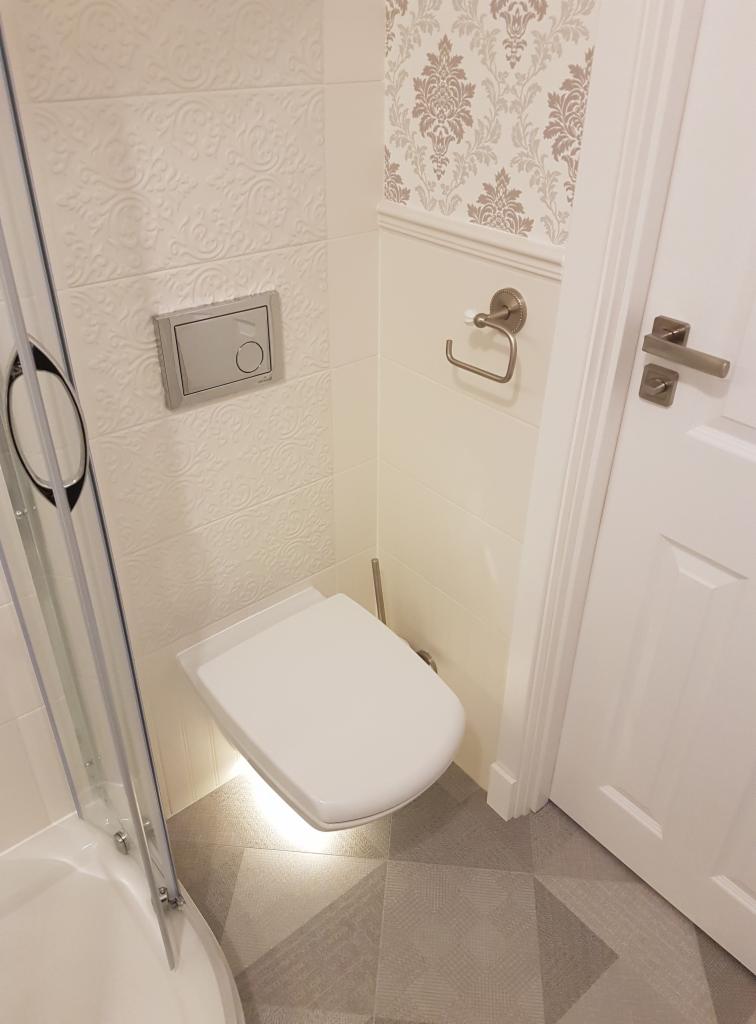 Nowoczesna łazienka z podwieszaną toaletą, podświetleniem LED, wzorzystymi płytkami i tapetą w kwiaty.