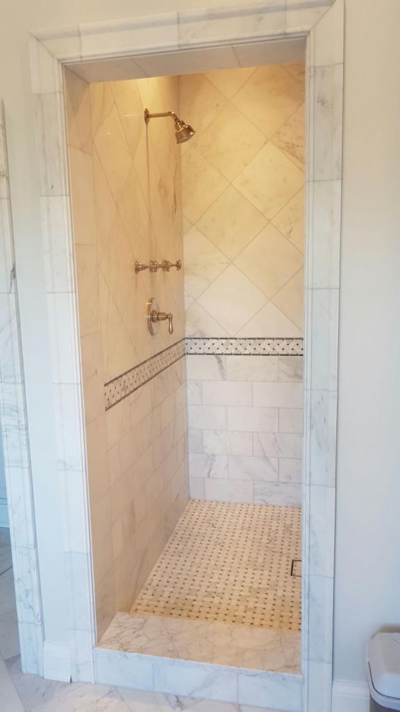 Wykończony prysznic z marmurowymi płytkami w różnych wzorach, w tym romby i prostokąty, z ozdobnym pasem płytek z małymi, ciemnymi punktami, oraz złotą armaturą.